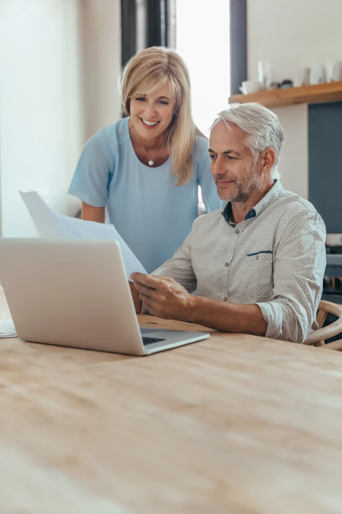 Smiling mature reading through investment documents and paying bills while doing their online banking at their kitchen table