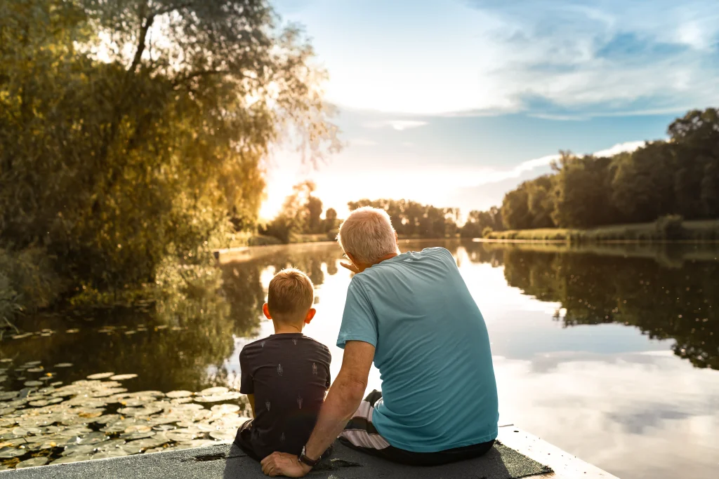 Grandfather and grandson sitting on a dock by a lake watching beautiful sunset.