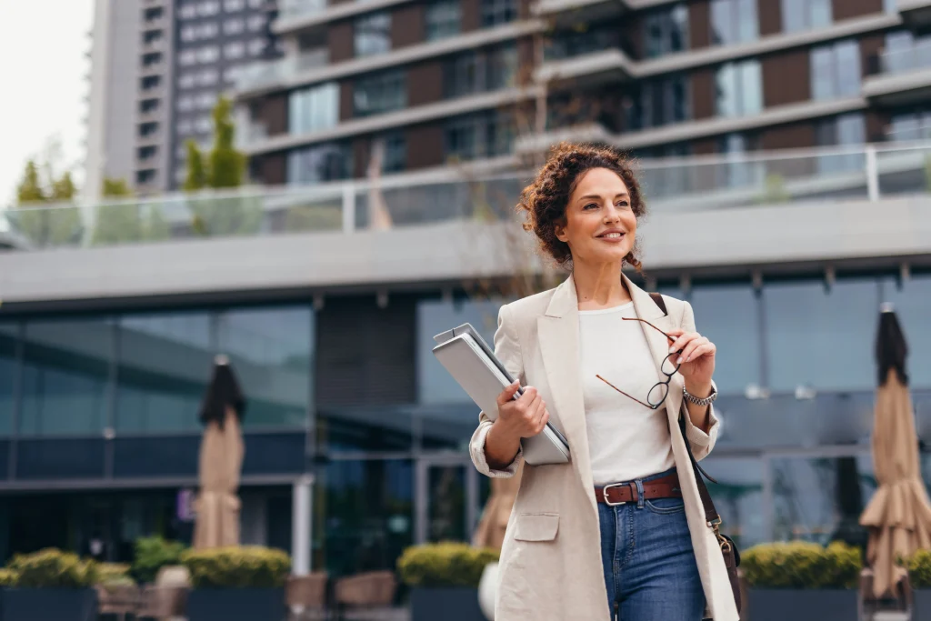 Smiling businesswoman walking confidently near modern office buildings, holding a laptop and eyeglasses, enjoying a productive workday in the vibrant city atmosphere