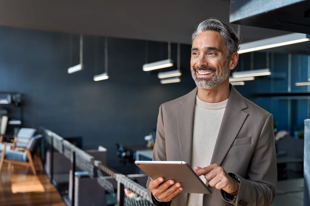 Happy confident mature business man executive standing in corporate office using digital tablet. Smiling middle aged professional businessman manager wearing suit looking away at work in modern space.